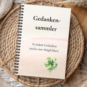 Könnte beinhalten: Ein spiralgebundenes Notizbuch mit dem deutschen Text "Gedankensammler" auf dem Cover, was "Gedankensammler" bedeutet. Das Cover hat ein Aquarelldesign mit einer Illustration eines vierblättrigen Kleeblatts. Das Notizbuch liegt auf einem geflochtenen Korb.