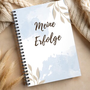 Könnte beinhalten: Ein spiralgebundenes Notizbuch mit hellblauem Aquarelldesign und den Worten "Meine Erfolge" in brauner Schrift. Das Cover zeigt Blattillustrationen und kleine Fußabdrücke. Das Notizbuch liegt auf einer beigen Oberfläche.
