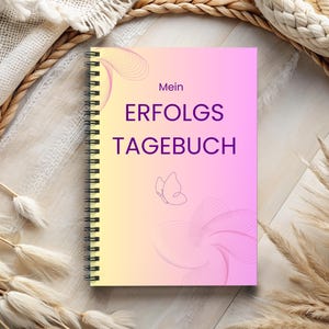 Könnte beinhalten: Ein spiralgebundenes Notizbuch mit einem gelb-rosa Farbverlauf. Auf dem Cover steht der Text "Mein ERFOLGS TAGEBUCH" in Lila, dazu ein Schmetterling und abstrakte Linien. Das Notizbuch liegt vor neutralem Hintergrund mit Pflanzen.