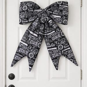 May include: A large, decorative bow made of black fabric with white text, hanging on a white door. The text includes words like "Jesus", "Prince", and "Peace". The bow is a festive decoration.