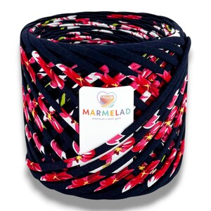 May include: A large roll of navy blue t-shirt yarn with a floral pattern of red and white flowers. The yarn is labeled "MARMELAD Premium t-shirt yarn". This yarn is suitable for various crafting projects.