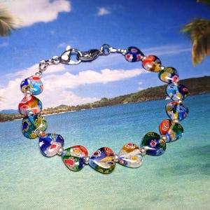 May include: A colorful bracelet featuring heart-shaped glass beads in shades of blue, red, green, and yellow. The beads are connected by a silver chain and clasp. The bracelet is displayed against a tropical background.