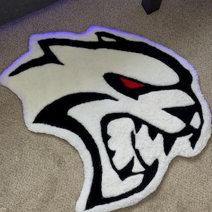 May include: A white and black rug shaped like a snarling cat's head with red eyes. The rug is made of a soft, textured material and is placed on a neutral-colored carpet.