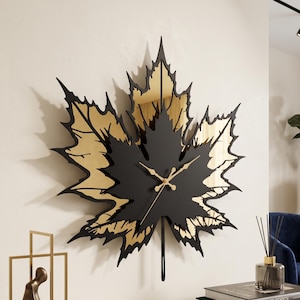 Pode incluir: Um relógio de parede em forma de uma grande folha de bordo, apresentando uma camada frontal em preto mate e uma camada posterior em tom dourado refletor. O relógio tem ponteiros dourados e um pêndulo fino pendurado no centro inferior.