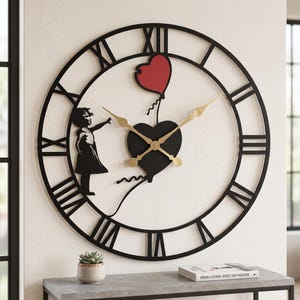 Puede incluir: Un reloj de pared circular grande con marco de metal negro y números romanos. El centro presenta una forma de corazón negro, un globo rojo en forma de corazón y la silueta de un niño extendiendo la mano. Manecillas de color dorado.
