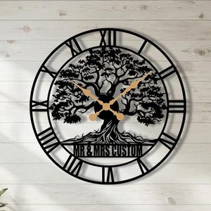May include: Black metal wall clock featuring a tree design with Roman numerals. The clock hands are gold. The text "MR & MRS CUSTOM" is displayed at the bottom of the tree. The clock is mounted on a white wooden wall.