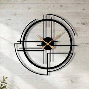 Könnte beinhalten: Eine große, schwarze, geometrische Wanduhr mit goldfarbenen Zeigern und einem schwarzen Mittelpunkt. Die Uhr ist an einer weißen Holzwand in einem Wohnzimmer befestigt. Das Design der Uhr zeigt sich kreuzende Linien und eine kreisförmige Form.