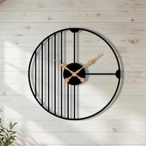 May include: A large, modern black metal wall clock with a circular design. The clock features a black face with gold-colored hands and a unique striped pattern on the left side. The clock is mounted on a white wooden wall in a living room setting.