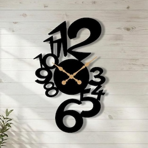 Peut inclure: Une horloge murale noire et moderne au design unique. L'horloge présente de grands chiffres noirs stylisés et des aiguilles dorées sur un cadran circulaire noir. Elle est fixée sur un mur en bois blanc, ajoutant une touche contemporaine à la pièce.