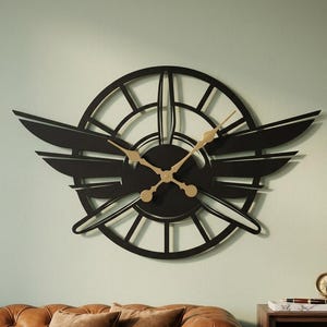 Könnte beinhalten: Eine große, kreisförmige schwarze Wanduhr mit einem stilisierten Propellerdesign und horizontalen Flügeln an den Seiten. Das Zifferblatt hat einen skelettierten Rahmen und goldfarbene Zeiger, die am Propellernabenpunkt befestigt sind.