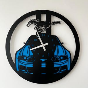 May include: Black and blue wall clock featuring a car silhouette with a running horse emblem. The clock has a circular design with a blue car outline and white clock hands. The numbers are in a blue font.