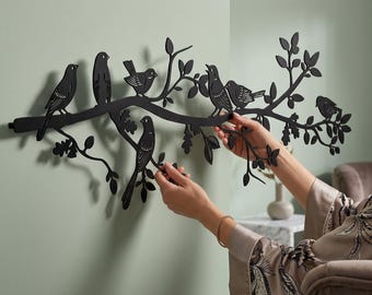May include: A matte black metal wall art piece featuring a horizontal tree branch silhouette with multiple birds perched among leaves and twigs. The design is elongated, showcasing detailed cutouts of avian figures and foliage extending across the wall.