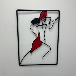 May include: Black metal wall art depicting a couple dancing. The woman is wearing a red dress and the man is wearing a red hat. The art is framed in a black square.