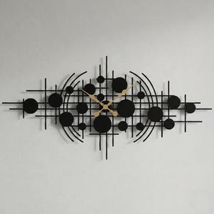 Op de afbeelding: Zwarte wandklok met een modern ontwerp. De klok heeft een reeks zwarte cirkels en lijnen, met goudkleurige wijzers. De klok is gemonteerd op een witte muur. Het ontwerp is abstract.
