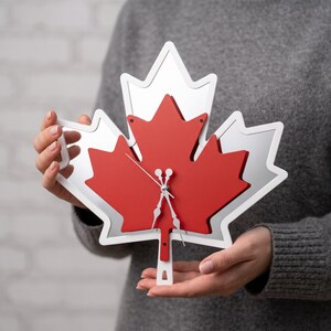 May include: A maple leaf-shaped wall clock featuring a layered design with a reflective mirror-finish back layer and a solid red front layer. The clock has white edges and thin, silver-colored metal hands positioned at the center of the leaf.