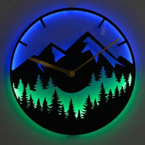 Op de afbeelding: Een ronde wandklok met een berg- en bos-silhouetontwerp. De klok heeft een zwarte wijzerplaat met goudkleurige wijzers en een blauw-groene LED-lichtrand. Het ontwerp van de klok omvat een bergketen en een bos van bomen.