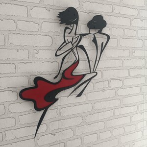 May include: A black metal wall sculpture depicting two stylized figures in a dancing pose. The taller figure features a flowing red dress element, while the shorter figure is rendered in solid black lines. The piece is mounted with visible screws.