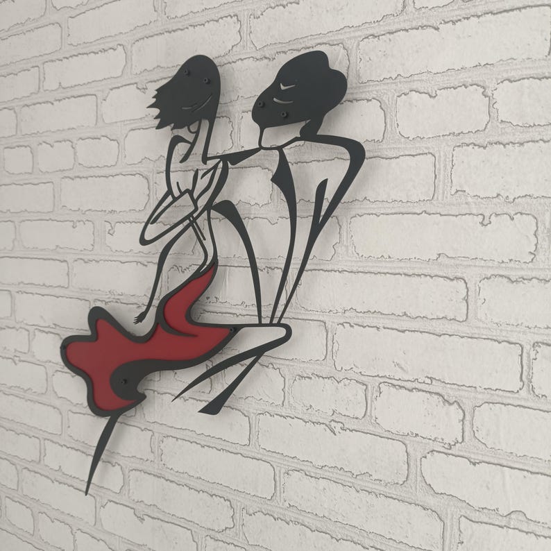 May include: A black metal wall art piece depicting two stylized figures in a dancing pose. The design features thin, continuous black lines forming the silhouettes of the figures, with one figure wearing a solid red skirt or dress element.