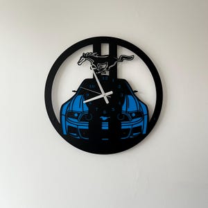 May include: Black and blue car-themed wall clock. The clock features a silhouette of a car with blue accents and a horse emblem. The clock hands are white, and the clock is mounted on a white wall.