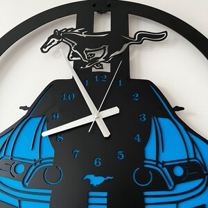 May include: Black and blue wall clock featuring a running horse silhouette. The clock has a car-shaped design with blue accents and silver clock hands. The numbers are in a blue font. The clock is a decorative item.