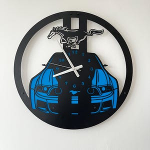 May include: Black and blue wall clock featuring a car design. The clock has a circular frame with a silhouette of a running horse at the top. The car's front is blue, and the clock hands are white.