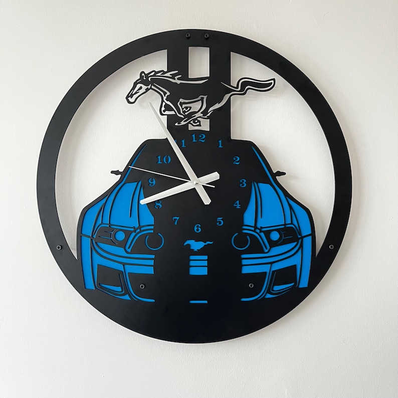 May include: Black and blue wall clock featuring a car design. The clock has a circular frame with a silhouette of a running horse at the top. The car's front is blue, and the clock hands are white.