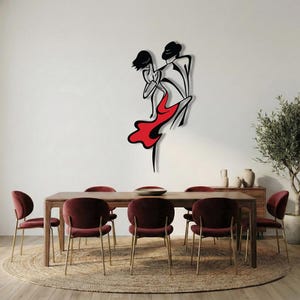 May include: A black metal wall art piece depicting a stylized dancing couple. The design features clean, minimalist lines with the dancer wearing a flowing red dress. The artwork is mounted on a plain white wall above a wooden dining table.