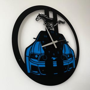 May include: Black and blue wall clock featuring a car silhouette with a running horse emblem. The clock has a circular black frame and silver hands. The car design is blue, and the horse is black.