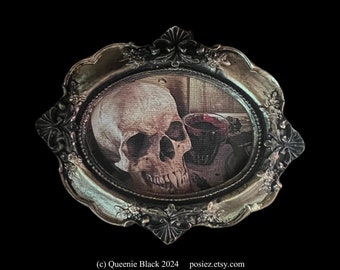 May include: An oval-shaped, antique-style frame with a skull print. The frame has ornate detailing and a silver finish. The skull is depicted in a realistic style, with a dark background. The image is a gothic or macabre art piece.