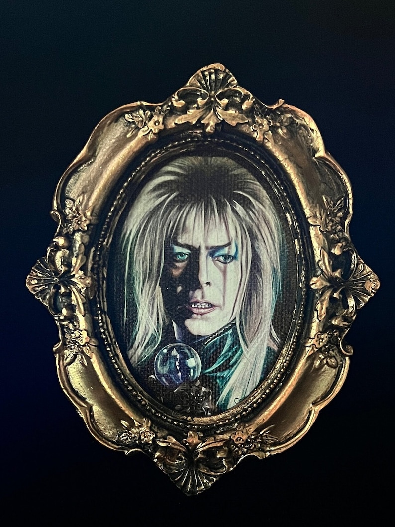 May include: A print of a Jareth from a labyrinth. The painting is framed in a gold oval frame with ornate details.