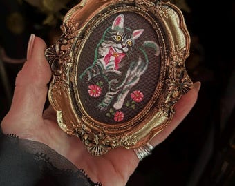 May include: A framed embroidered artwork featuring a cat with a red bow and pink ears. The cat is gray and black, set against a dark background with pink flowers. The frame is ornate and gold-colored.