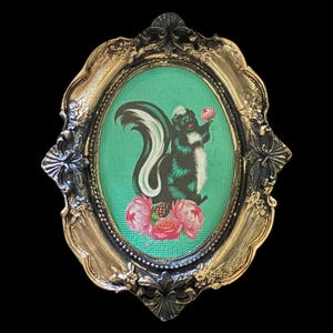 May include: A framed illustration of a black and white skunk holding a pink flower. The skunk is surrounded by pink flowers and green foliage. The frame is gold with black accents.