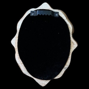 May include: A gold-colored oval-shaped frame with a black velvet backing. The frame has a sawtooth hanger on the back for hanging on a wall.