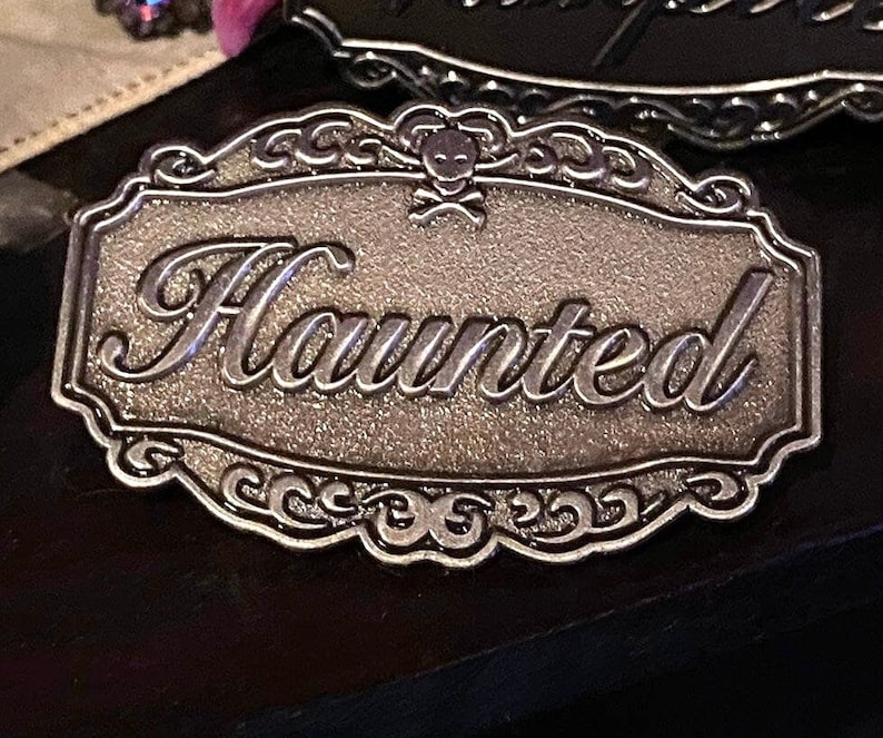 May include: A silver-colored metal sign with an ornate border and the word "Haunted" in a decorative script. A skull and crossbones design is above the word.