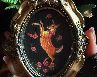 Puede incluir: Un marco ovalado dorado con un diseño ornamentado rodea una pequeña pintura de un ciervo bailando. El ciervo es naranja con una pajarita rosa, sobre un fondo negro con flores rosas. El marco es sostenido por una persona.
