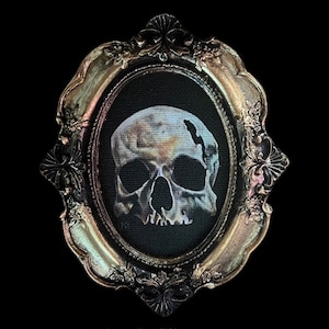 May include: A white skull with a crack in the top of the head is painted on a black canvas. The canvas is framed in a gold ornate oval frame.