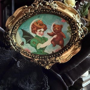 May include: An oval-shaped, antique gold-colored frame with a whimsical illustration. The artwork depicts a woman in a green outfit and a brown teddy bear in a bubble. The background is a light blue with floral accents. The frame is held by a black gloved hand.