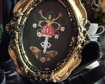 May include: An oval, gold-framed artwork featuring a black background with a floral and bat design. The artwork has a gold ornate frame. The floral design is red, pink, and yellow. The artwork is held by a black gloved hand.