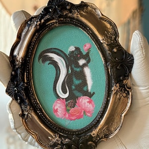 May include: A framed cross-stitch of a black and white skunk holding a pink flower. The skunk is surrounded by pink flowers and green leaves. The frame is oval shaped and has a gold finish.