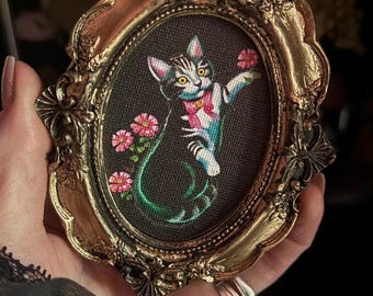 May include: A framed embroidery of a white and gray tabby cat with pink flowers and a green vine. The cat is wearing a pink bow tie and is reaching up with its paw. The frame is gold and oval shaped.