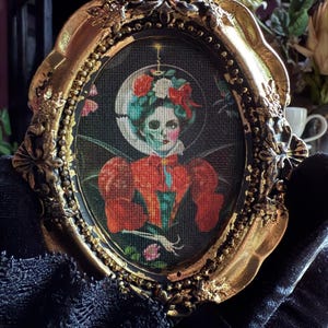 May include: An oval-shaped, ornate gold frame surrounds a gothic-style portrait. The artwork depicts a woman with a skull face, adorned with flowers and a red bow. She wears a red dress with puffed sleeves. The background is dark, with a crescent moon.