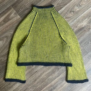 Puede incluir: Un suéter de manga larga con cuello alto y un patrón de punto acanalado vertical. La prenda es principalmente de color verde lima con ribetes azul marino oscuro a lo largo del cuello, el dobladillo y los bordes de las mangas anchas.