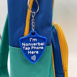 May include: A blue shield-shaped keychain with the text "I'm Nonverbal Tap Phone Here" and a sound wave symbol. The keychain hangs from a silver chain and ring attached to a blue, green, and yellow backpack.