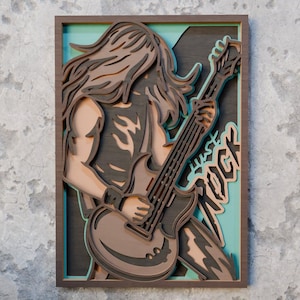 Può includere: Un'opera d'arte murale in legno a strati raffigurante un chitarrista con i capelli lunghi. L'opera presenta una cornice marrone, uno sfondo turchese e le parole "Just Rock". Il musicista è in tonalità di marrone e grigio.