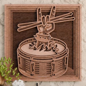 May include: Wooden wall art featuring a layered design of a drum set with a hand making the rock and roll sign. The art piece is framed in a square, with a light brown and dark brown color scheme. The drumsticks are visible.