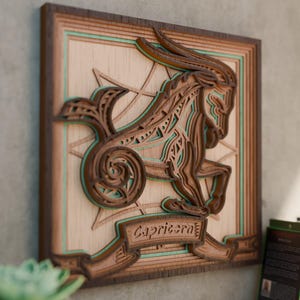 May include: Wooden Capricorn zodiac sign wall art. The layered design features a brown goat figure with teal accents, set against a light wood background. The sign's name is displayed on a banner. The artwork is framed in a dark brown border.