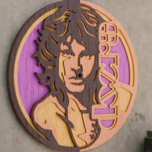 Könnte beinhalten: Runde Wandkunst aus Holz mit einem Porträt eines Mannes mit langen Haaren. Das Design enthält den Namen der Band in einer stilisierten Schriftart. Das Kunstwerk verwendet Braun-, Lila- und Gelbtöne und erzeugt einen geschichteten Effekt. Es ist an einer grauen Wand befestigt.