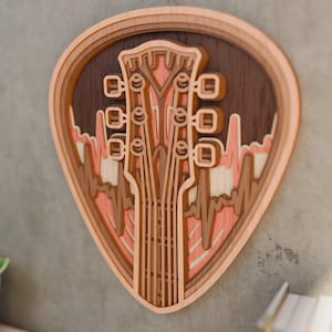 Op de afbeelding: Een gelaagd houten wandkunstwerk in de vorm van een plectrum. Het ontwerp toont een gitaarkop en een geluidsgolfpatroon in bruin, perzik en crème. Het kunstwerk is op een grijze muur gemonteerd.