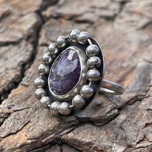 May include: A silver ring with a purple gemstone set in a bezel setting. The ring is surrounded by a circle of silver beads.
