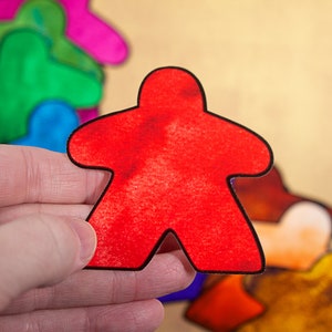 May include: A red meeple game piece, a common element in board games, is held in a hand. The meeple is made of a textured material and has a black outline.